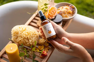 Gentle body products like rose and sandalwood ritual spray held over a spa-inspired bath tray with flowers and candles.