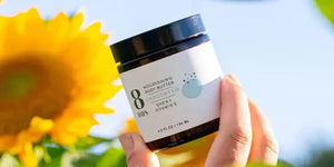 Natural body butter jar labeled "8 Days Nourishing Body Butter Unscented" with shea and vitamin E, held against a sunflower backdrop