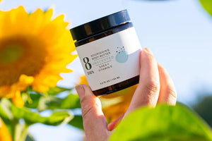 Natural herbal skincare body butter with shea and vitamin E, held against a backdrop of vibrant sunflowers in sunlight.