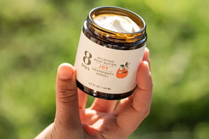 A hand holding a nourishing body butter jar with grapefruit and neroli against a green blurred background